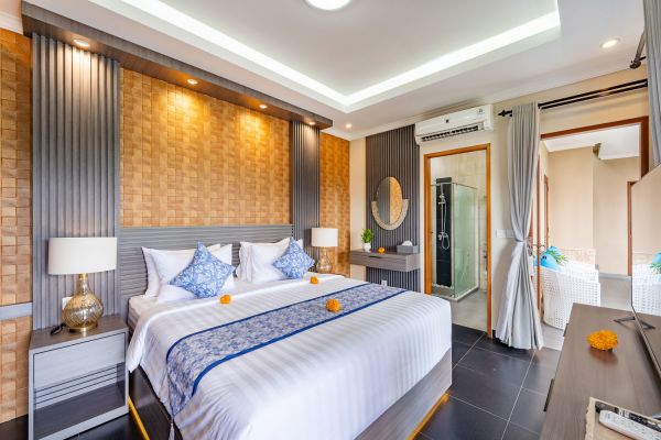 Spacious Balinese-style luxury bedroom with king bed, blue bedding, frangipani flowers, wooden walls, en-suite shower, and modern tropical decor.